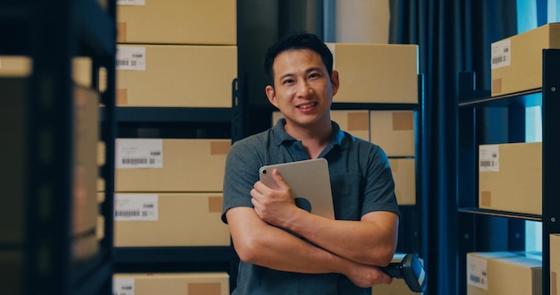 Un homme d'affaires asiatique prospère se tient devant une étagère sur une tablette numérique et un lecteur de codes-barres souriant et regardant la caméra fier des affaires dans l'entrepôt la nuit Concept de petite entreprise de démarrage