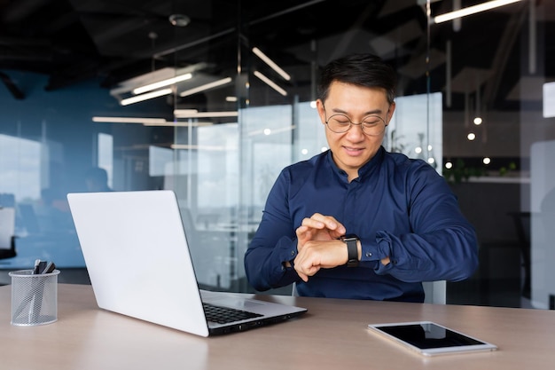 Homme d'affaires asiatique prospère utilise une montre intelligente sur le lieu de travail, l'homme choisit des applications et sourit heureux avec les applications