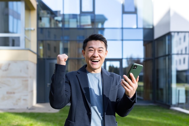 Homme d'affaires asiatique sorti célébrant la réussite en regardant un smartphone en plein air Homme d'affaires prospère célébrant la victoire avec un téléphone portable à l'extérieur