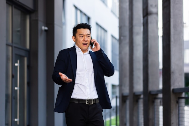 Un homme d'affaires asiatique à succès en colère et sérieux explique des informations aux employés par téléphone, parle près du bureau à l'extérieur