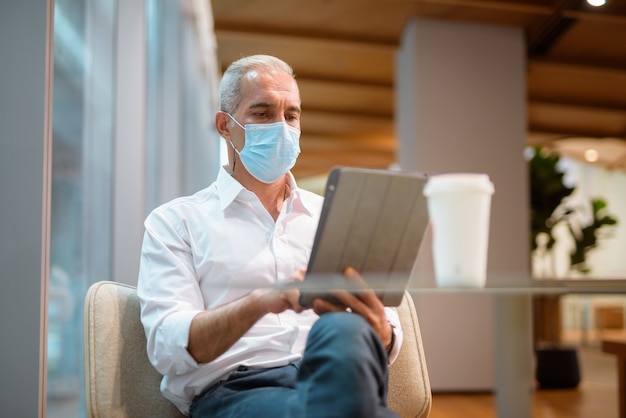 Homme d'affaires assis au café et utilisant une tablette numérique tout en portant un masque facial et un tir horizontal de distanciation sociale