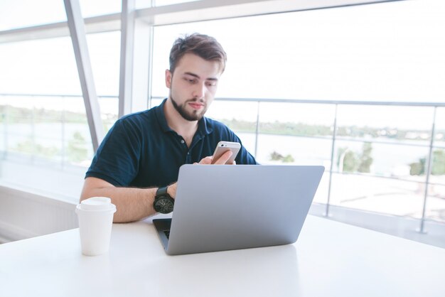 Homme d'affaires assis dans un coworking à une fenêtre avec un ordinateur portable et un café et utilise un smartphone