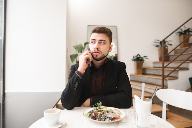 Homme d'affaires assis dans un restaurant avec salade et une tasse de café sur la table, parler sur le smartphone