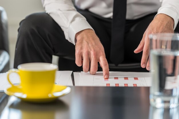 Homme d'affaires assis à une table de club dans son bureau examinant des tableaux statistiques dans son rapport avec une tasse de café jaune et un verre d'eau sur la table