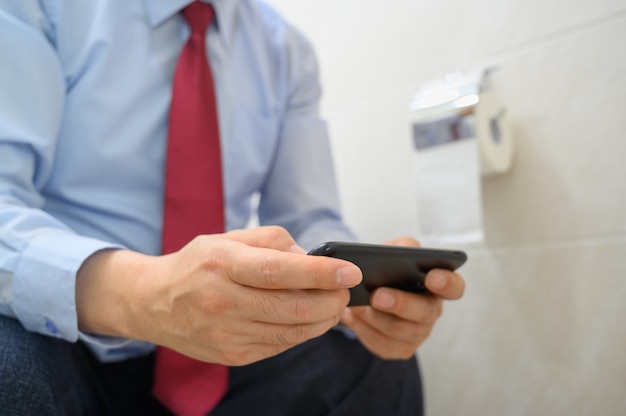 Homme d'affaires assis sur les toilettes dans la salle de bain et à l'aide d'un smartphone.