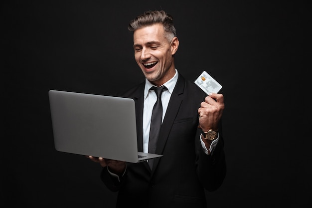 Homme d'affaires attrayant et confiant portant un costume isolé sur un mur noir, utilisant un ordinateur portable, montrant une carte de crédit en plastique