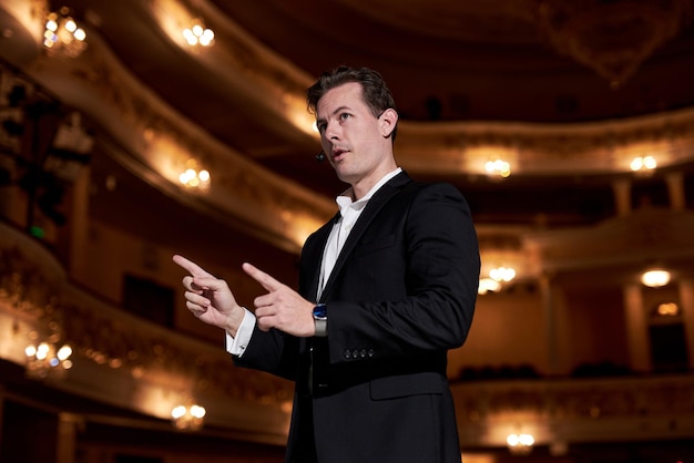 Homme d'affaires attrayant debout sur scène. Concept d'étoile, de spectacle, de discours et de présentation. Beau présentateur caucasien parlant au public dans l'auditorium de la salle de conférence, faisant des gestes.