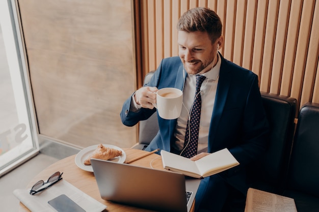 Homme d'affaires attrayant à distance après une réunion financière en ligne avec une tasse de café au lait à la main dans un café et satisfait des résultats, utilisant un ordinateur portable avec des écouteurs tout en vérifiant les entrées dans le classeur