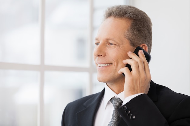 Homme d'affaires au téléphone. Portrait d'un homme mûr gai en tenue de soirée parlant au téléphone et souriant tout en se tenant près de la fenêtre