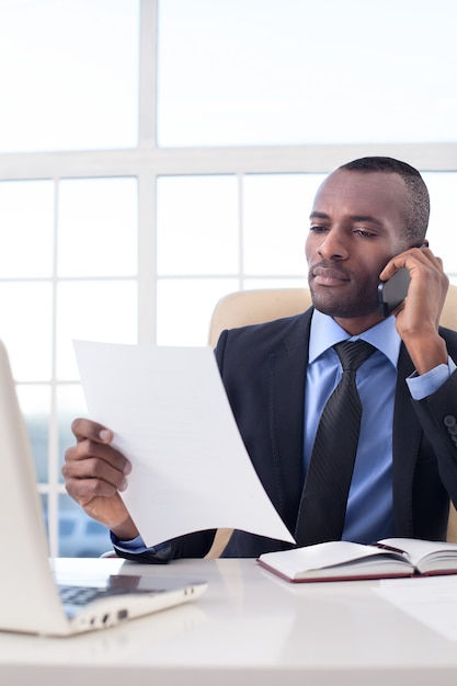 Homme d'affaires au travail. Jeune homme d'affaires africain confiant parlant au téléphone portable et vérifiant des documents alors qu'il était assis sur son lieu de travail