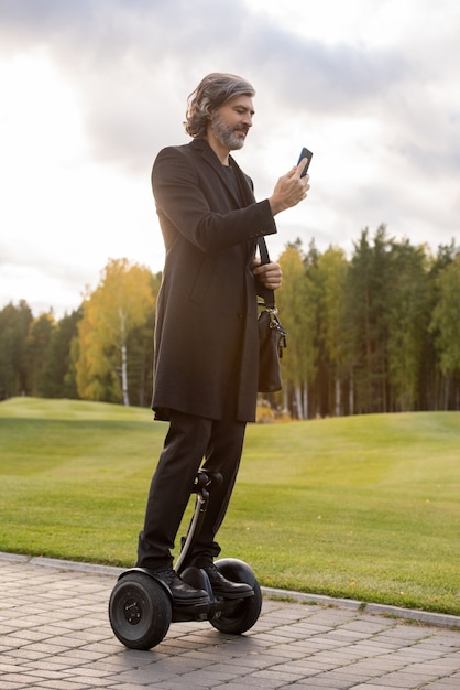 Photo homme d'affaires aux cheveux gris barbu en manteau noir utilisant un smartphone en se tenant debout sur un gyroscope contre une grande pelouse verte et des arbres