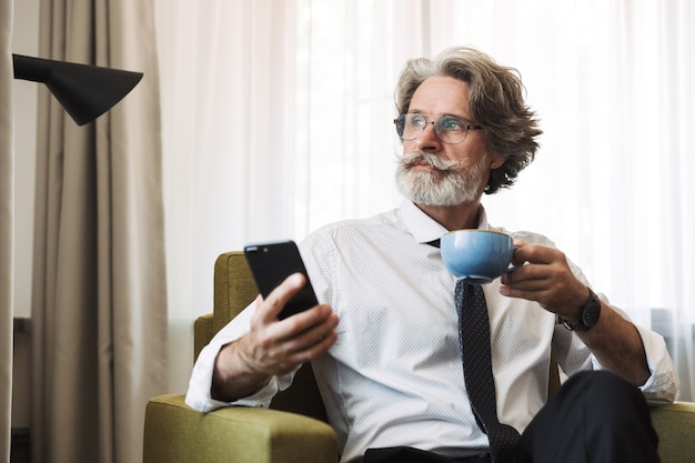 Homme d'affaires aux cheveux gris concentré, pensant sérieusement, assis sur une chaise à l'intérieur à la maison à l'aide d'un smartphone buvant du café.