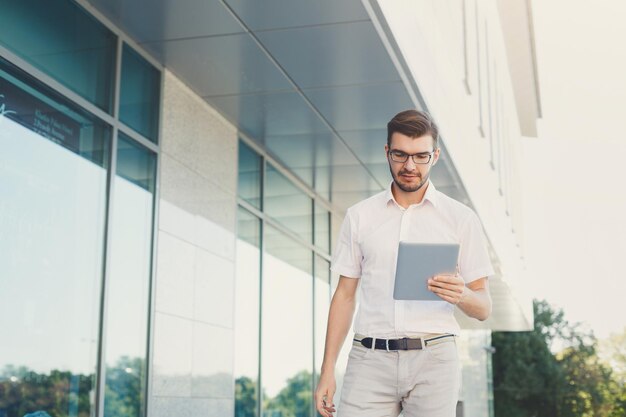 Un homme d'affaires ou un avocat concentré attrayant dans des lunettes lit sur une tablette numérique tout en se tenant près d'un immeuble de bureaux moderne, d'un recadrage, d'un espace de copie. Concept commercial et technologique