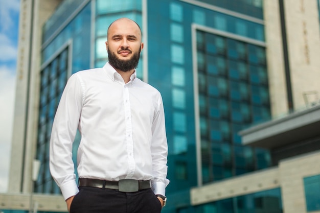 Homme d'affaires avec barbe en chemise blanche debout près d'un immeuble de bureaux et regardant à huis clos