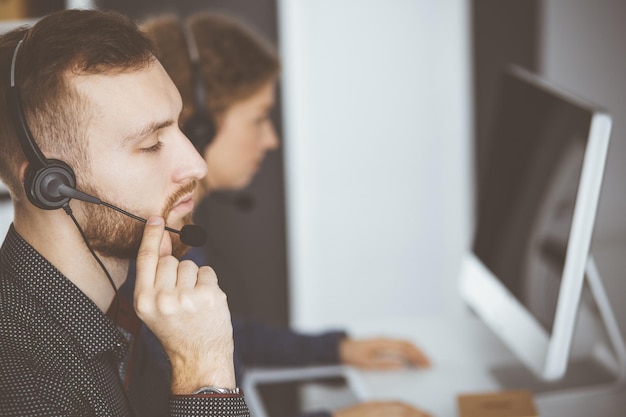 Homme d'affaires à barbe rousse parlant par casque près de sa collègue féminine alors qu'il était assis dans un bureau moderne. Groupe de personnes diverses dans le centre d'appels. Télémarketing et service à la clientèle.