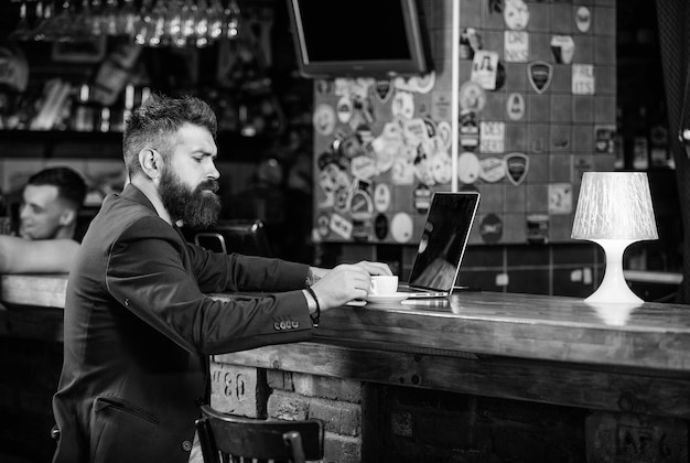 Homme d'affaires barbu assis dans un pub avec un ordinateur portable et une tasse de café Le directeur travaille en ligne tout en savourant un café