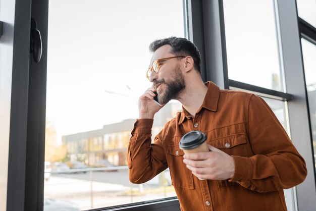 un homme d'affaires barbu satisfait avec des lunettes à la mode et une chemise élégante tenant une tasse de papier pendant