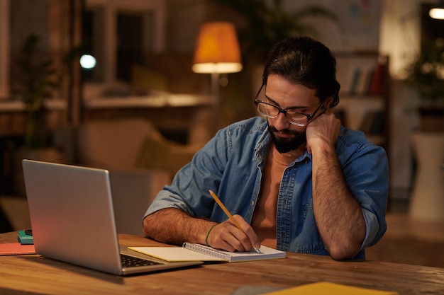 Homme d'affaires barbu sérieux, prendre des notes dans un bloc-notes assis sur son lieu de travail devant un ordinateur portable