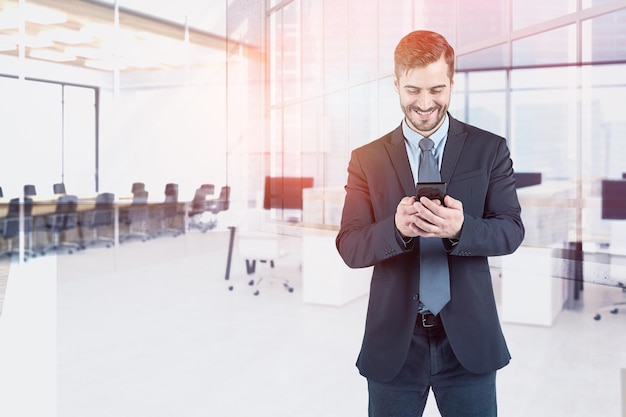 Photo un homme d'affaires barbu souriant regarde son smartphone dans un bureau moderne avec une double exposition du gratte-ciel.