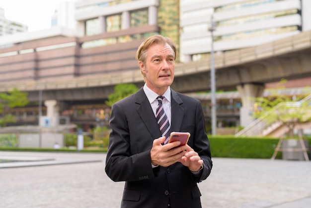 Homme d'affaires beau mature pensant tout en utilisant le téléphone dans la ville à l'extérieur