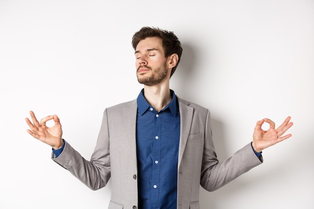 Homme d'affaires calme et concentré, méditant les yeux fermés et les mains écartées sur le côté, trouvant la paix dans la méditation, pratiquez la respiration du yoga, debout sur fond blanc.