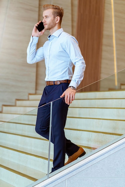 Un homme d'affaires caucasien intelligent qui a réussi à marcher dans l'escalier permet à la communication avec un smartphone de communiquer avec bonheur et un arrière-plan de bureau moderne enthousiaste