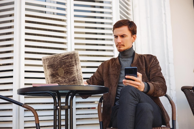 Homme d'affaires concentré utilisant un smartphone et un ordinateur portable tout en travaillant en ligne sur un projet dans un café de la ville