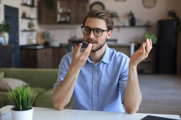 Homme d'affaires confiant dans des verres tenant un smartphone près de la bouche pour enregistrer un message vocal ou activer l'assistant numérique.