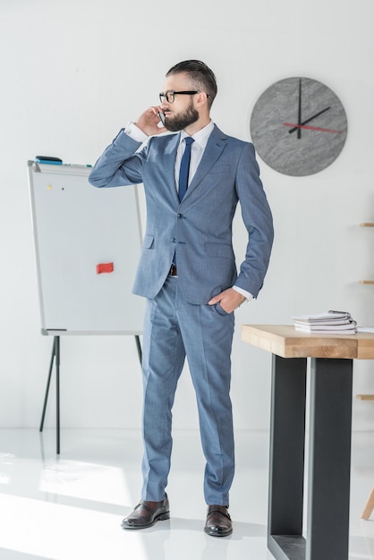 Homme d'affaires confiant parlant sur smartphone tout en se tenant sur le lieu de travail au bureau