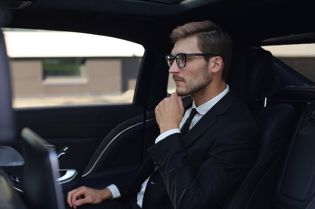 Homme d'affaires confiant et réfléchi gardant la main sur les lunettes alors qu'il était assis dans la voiture de luxe.