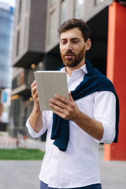Homme d'affaires confiant en tenue formelle debout près d'un immeuble de bureaux et utilisant une tablette numérique pour une conversation vidéo. Concept de personnes et de technologie moderne.