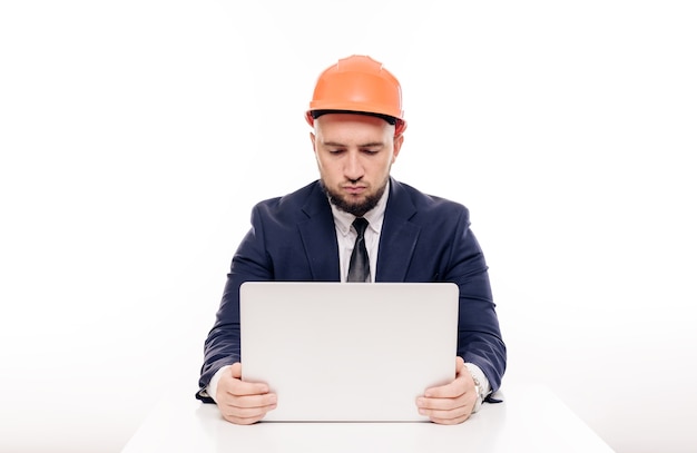 Un homme d'affaires constructeur fatigué dans un casque orange regarde l'écran de l'ordinateur portable et étudie le projet de construction. S'assoit à table et boit du café, nerveux