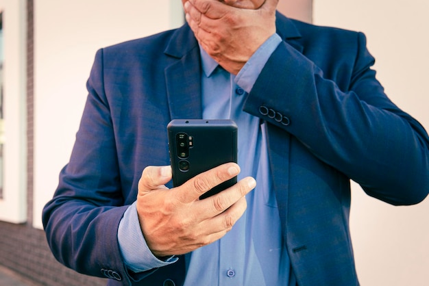 Un homme d'affaires en costume bleu regarde un smartphone dans sa main Le concept de communication en ligne Un homme surpris tient un téléphone et lit un message pointant dessus avec le doigt