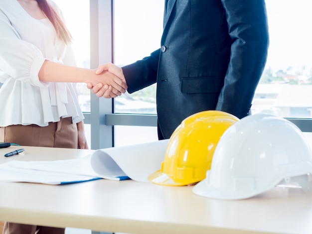 Homme d'affaires en costume, ingénierie ou architecte et femme se serrant la main sur le plan et le casque de sécurité jaune et blanc sur le bureau sur la fenêtre en verre.