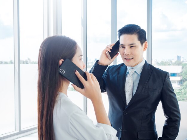 Homme d'affaires en costume et jeune femme appelant avec un téléphone portable sur une immense vitre en milieu de travail dans un immeuble élevé.