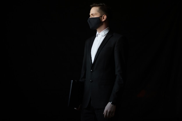 Homme d'affaires en costume noir portant un masque de protection tient un ordinateur portable