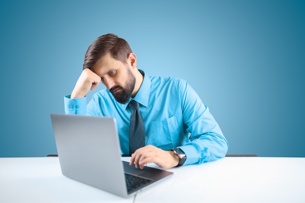 Un homme d'affaires dans une chemise bleue et une cravate travaille pensivement à un ordinateur sur un projet responsable, l'homme a appuyé sa main sur sa tête