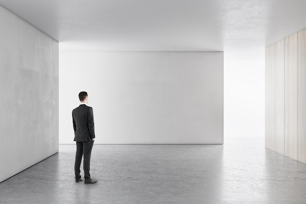 Homme d'affaires dans un intérieur en béton clair avec un mur vide regardant un lieu de maquette pour votre publicité