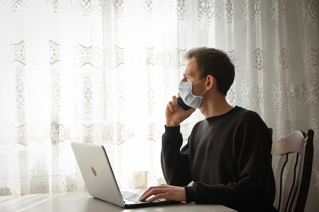 Homme d'affaires dans un masque médical et dans un peignoir à domicile pendant les travaux de quarantaine au café. Un jeune homme dans un café travaille à un ordinateur portable près de la fenêtre. Isolement, coronavirus, travail à domicile.