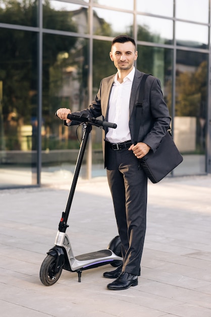 Homme d'affaires debout devant la ville tenant un scooter électrique et regardant la caméra urbaine moderne