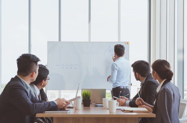 Homme d&#39;affaires dirigeant écrit sur le tableau blanc présente le graphique de marketing d&#39;entreprise lors d&#39;une réunion avec des collègues de bureau. Présentation de réunion d&#39;équipe d&#39;entreprise, Conférence Business Planning Concept