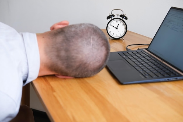 Homme d'affaires dormant sur le bureau à côté d'un ordinateur portable et d'un réveil