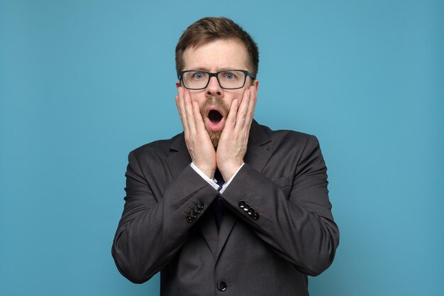 Un homme d'affaires effrayé en costume tient les mains sur son visage en état de choc avec de grands yeux ouvrant la bouche