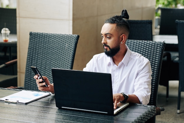 Homme d'affaires élégant avec chignon homme travaillant sur un ordinateur portable à la table du café et lisant des messages texte d'un client ou d'un collègue