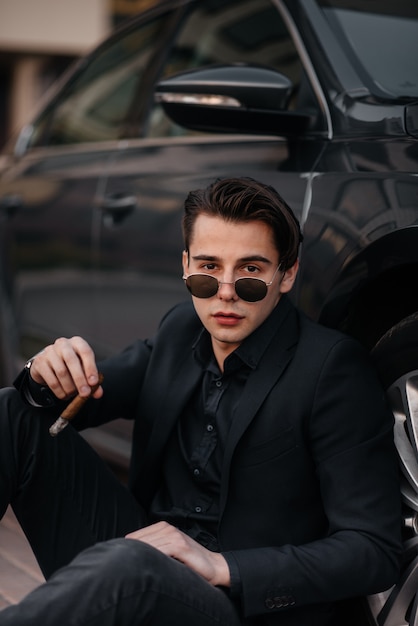 Un homme d'affaires élégant fume des cigares près d'une voiture de luxe. Mode et affaires.