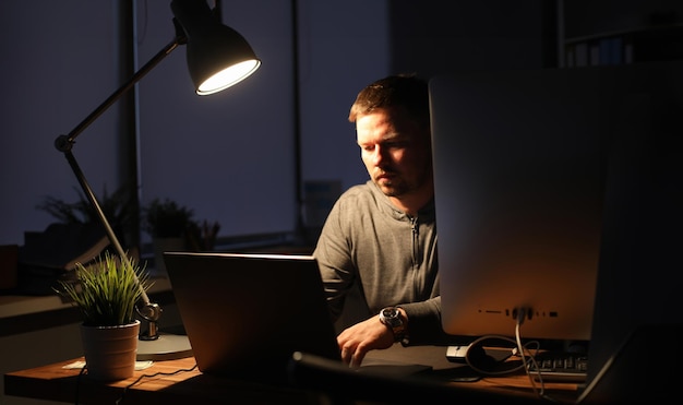 L'homme d'affaires est assis dans un bureau le soir à table à la lumière d'une lampe Utilise un ordinateur portable et un PC L'écrivain remplit les frais Concept de démarrage d'entreprise