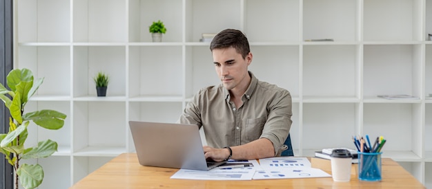 Un homme d'affaires est assis dans son bureau avec un ordinateur portable et de la papeterie, sur son bureau il y a des documents financiers qu'il vérifie, il tape, envoie des SMS, parle à son partenaire via un messager pour ordinateur portable.