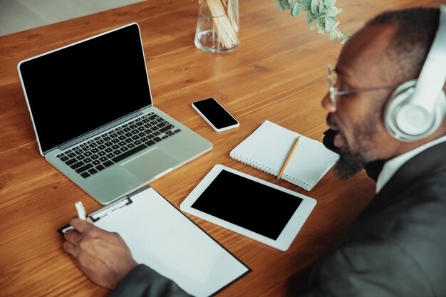 Homme d'affaires ou étudiant travaillant à domicile isolé ou maintenu en quarantaine à cause du coronavirus. Homme afro-américain utilisant un ordinateur portable, une tablette et un casque. Conférence en ligne, cours, bureau à distance.