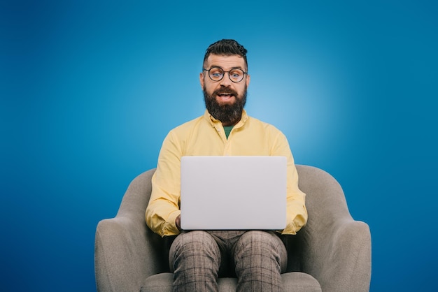 Homme d'affaires excité utilisant un ordinateur portable assis dans un fauteuil isolé sur bleu