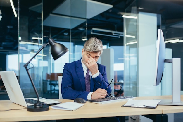 Un homme d'affaires expérimenté aux cheveux gris travaille à l'ordinateur jusqu'à ce qu'il soit fatigué tard et souffre de maux de tête graves, de stress et de dépression